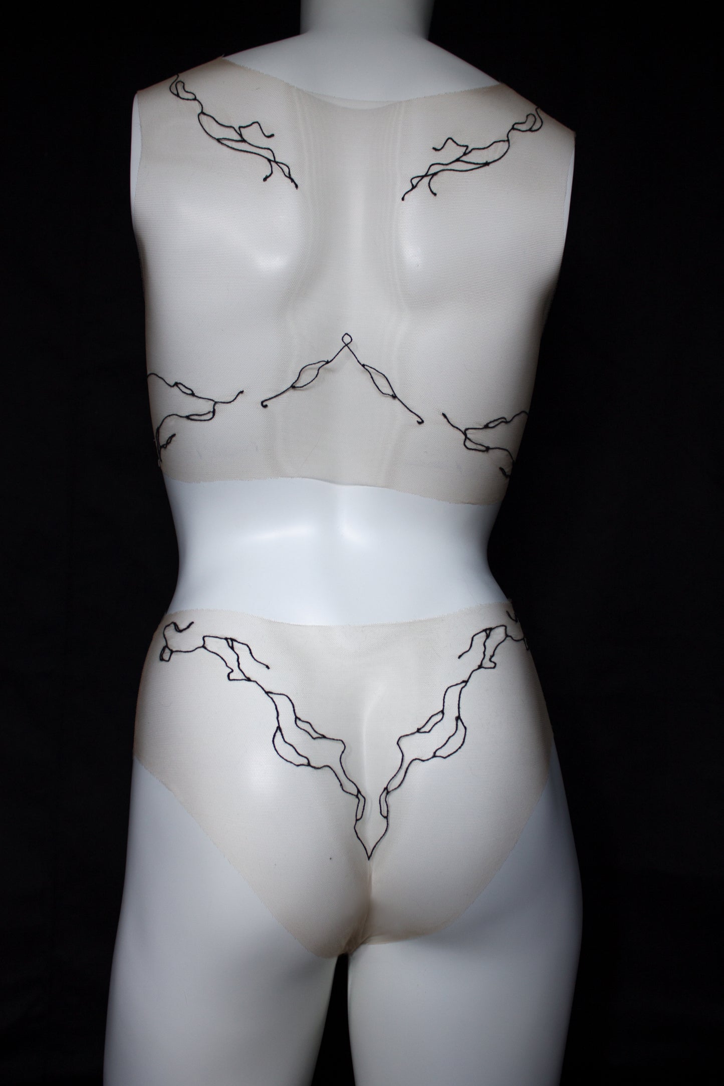 Mannequin wearing sheer beige garment with black lace details on a black background