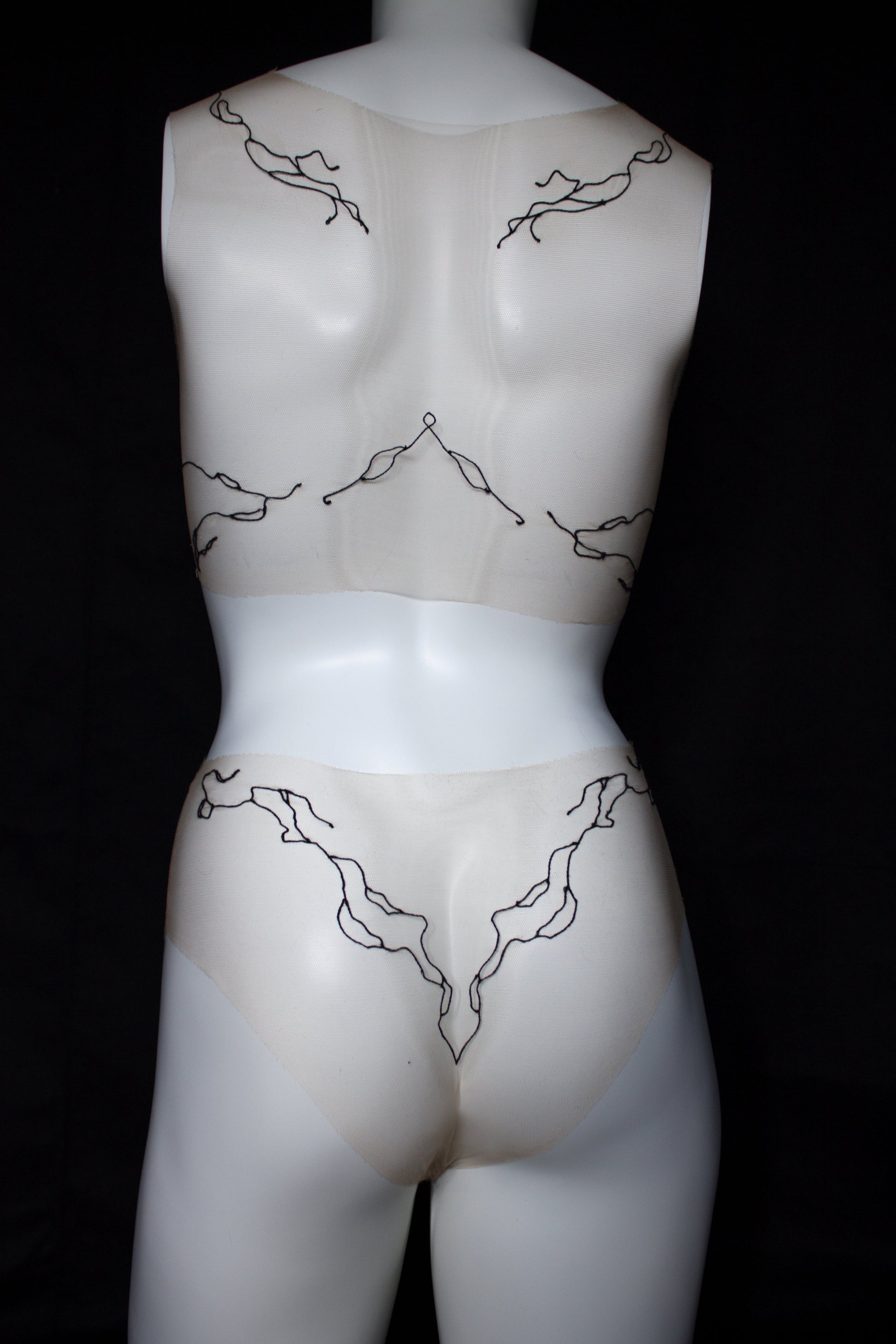 Mannequin wearing sheer beige garment with black lace details on a black background